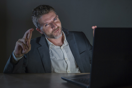 young excited and attractive businessman crossing fingers for luck hoping to win online bet while working late night at office laptop computer desk in internet gambling and money makingの写真素材