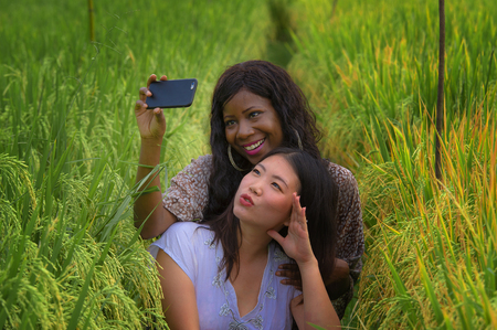 mixed ethnicity Asian Korean girl and black African American woman taking girlfriends selfie with mobile phone together isolated at rice field enjoying exotic tropical holidays in traditional villageの写真素材