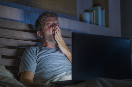 attractive tired and stressed workaholic man working late night exhausted on bed busy with laptop...