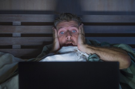 lifestyle portrait of young scared and stressed man in bed watching internet horror movie late night with  laptop computer or bedroom television in panic face expression covering with blanketの写真素材