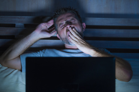 attractive tired and stressed workaholic man working late night exhausted on bed busy with laptop computer yawning feeling sleepy and overworked in business project deadline stress conceptの写真素材
