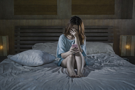 young depressed Asian Chinese girl using mobile phone crying on bed at night feeling sad and depressed victim of cyber bullying or broken heart suffering depression in desperate woman expressionの写真素材