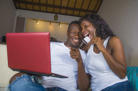 lifestyle portrait of young happy and attractive black African American couple enjoying at home using credit card and laptop computer for online shopping in internet banking electronic business conceptの写真素材