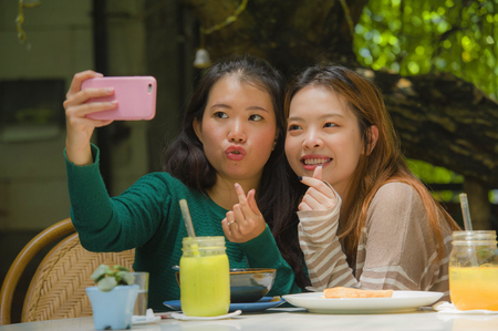 lifestyle portrait of two young happy and cute Asian Chinese girlfriends taking selfie portrait picture with mobile phone camera for using on internet social media while having healthy brunch at cafeの写真素材
