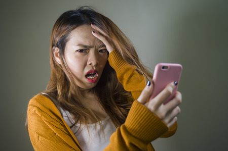 young cute and sad Asian Korean girl feeling broken heart and desperate holding mobile phone suffering relationship break up dumped via internet message isolated on dark backgroundの写真素材