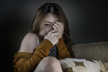 young attractive and cute scared Asian Korean woman alone at home living room couch watching horror movie or TV drama holding remote controller in fear and panic face expressionの写真素材