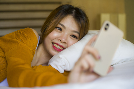 young attractive and relaxed Asian Chinese girl using mobile phone at home bedroom enjoying internet online dating app or social media lying on bed in teenager student lifestyle conceptの写真素材