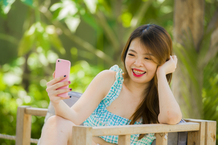 lifestyle portrait of young happy and attractive Chinese student girl at resort garden taking selfie photo with mobile phone camera enjoying holidays relaxed isolated in tropical vegetation backgroundの写真素材