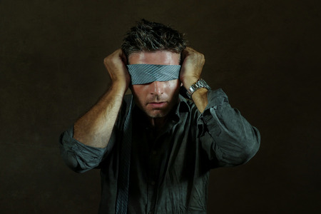 young lost and confused man blindfolded with necktie playing internet trend dangerous viral challenge with eyes blind acting doubtful guided by intuition isolated on dark backgroundの写真素材