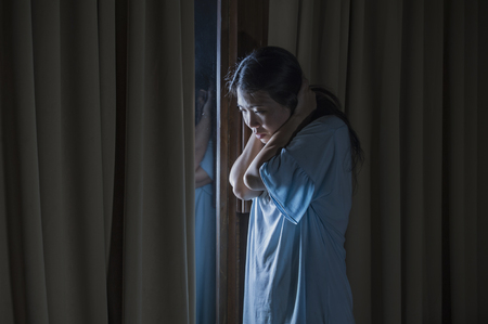 dramatic indoors portrait of young sad and depressed Asian Japanese woman crying desperate broken heart suffering depression and anxiety in dark light at home curtains window in sadness conceptの写真素材