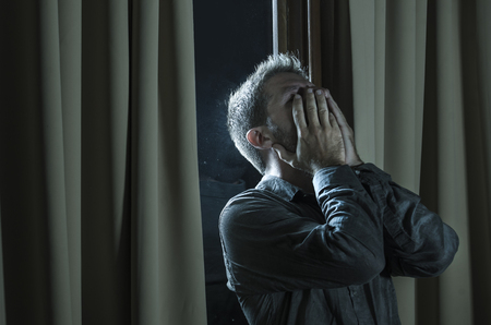 dramatic portrait of young handsome sad and depressed man desperate suffering depression problem feeling broken heart pain looking desperate at home by curtains window at nightの写真素材
