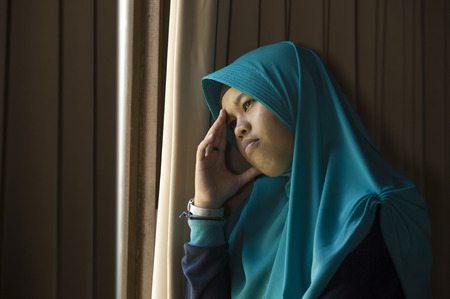 lifestyle portrait of young sad and depressed Muslim woman in Islam traditional Hijab head scarf at home window feeling unwell suffering depression crisis and anxiety problem thoughtful and helplessの写真素材