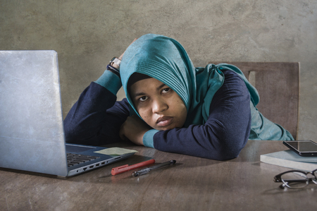 young stressed and overwhelmed Muslim student woman in Islam hijab head scarf  studying tired feeling overworked working with laptop computer and University books preparing examの写真素材