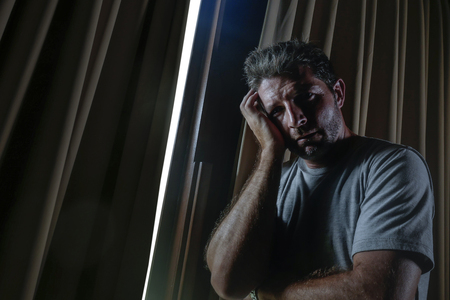 dramatic light indoors portrait of young sad and depressed attractive man at home looking through room window thoughtful and pensive lost in pain broken heart suffering depression problemの写真素材