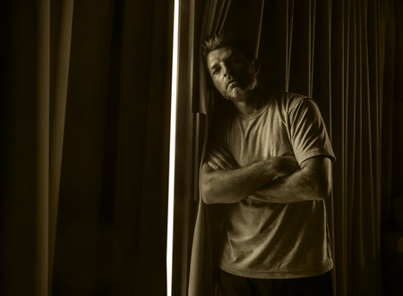 dramatic light indoors portrait of young sad and depressed attractive man at home looking through room window thoughtful and pensive lost in pain broken heart suffering depression problemの写真素材