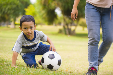 portrait in city park of happy Asian Indonesian mother playing football with little 5 years old son running together excited laughing having fun in soccer fan child and healthy lifestyle educationの写真素材