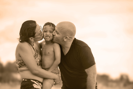 young happy and romantic mixed ethnicity couple Caucasian man and Asian Indonesian woman kissing their little son, the parents holding the kid and playing at tropical beach on holidays tripの写真素材