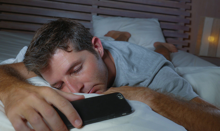 lifestyle portrait of young tired and exhausted man sleeping or falling asleep while networking in bed late at night in internet and social media smart phone addiction and overwork conceptの写真素材
