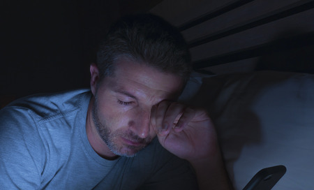 lifestyle portrait of young tired and sleepy man networking late at night lying on bed at home bedroom in the dark in internet and social media online app addiction conceptの写真素材