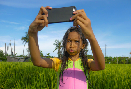 7 or 8 years old sweet and pretty female child outdoors at rice field landscape taking selfie portrait  photo with mobile phone camera enjoying holidays in children and internet technology conceptの写真素材