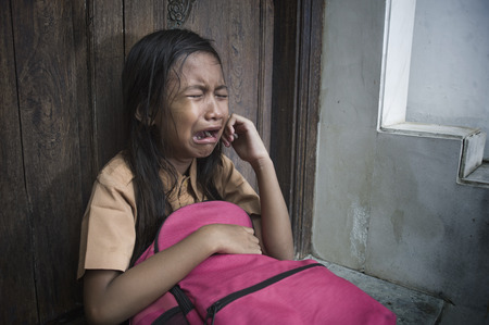 7 or 8 years child in school uniform sitting outdoors on the floor crying sad and depressed holding...