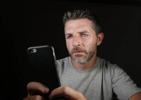 close up face portrait of young attractive and confident white man with blue eyes  using online dating app or internet social media on mobile phone isolated on black background in communication conceptの写真素材