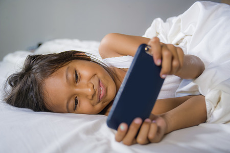 lifestyle portrait of sweet happy and beautiful 7 years old child having fun playing internet game with mobile phone lying on bed cheerful and excited in kid and domestic technology conceptの写真素材