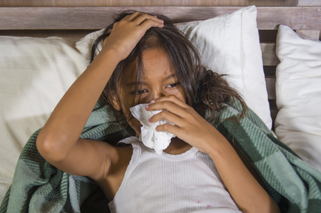 lifestyle home portrait of young beautiful and sweet 8 years old female child holding tissue paper sneezing her nose lying sick on bed having flu or cold virus feeling sad and unwellの写真素材