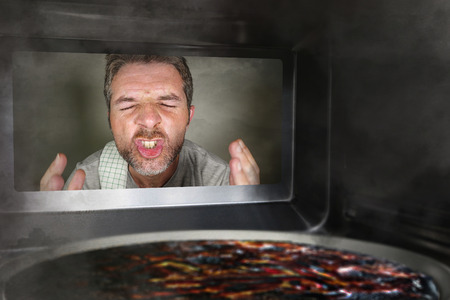 young messy and funny dummy man in the kitchen looking through microwave or oven pizza burning...