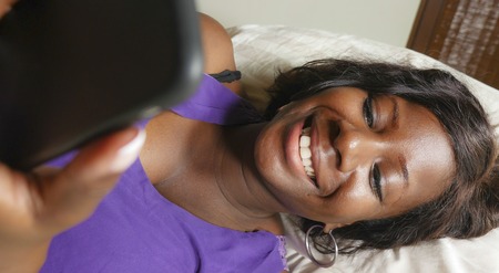 young beautiful and happy 30s black Afro American woman lying relaxed on bed using internet mobile phone smiling cheerful networking social media business or online dating at home bedroomの写真素材