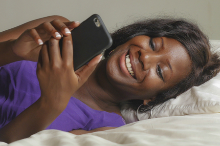 young beautiful and happy 30s black African American woman lying relaxed on bed using internet mobile phone smiling cheerful networking social media business or online dating at home bedroomの写真素材