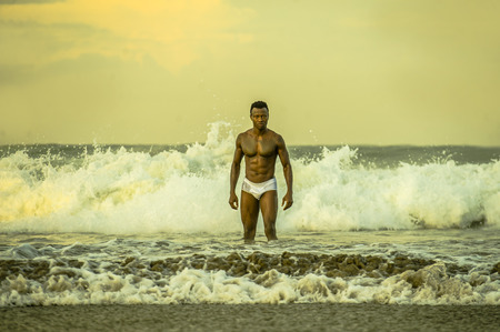 artistic portrait of young attractive and fit black afro American man with sexy muscular body and sixpack abs posing cool on beach with big sea waves and foam around in beauty and health conceptの写真素材