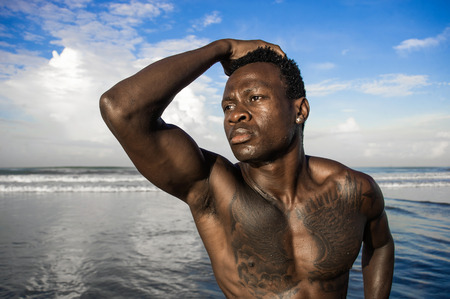 close up head and shoulders portrait of young attractive and fit black African American man with strong muscular body posing cool model attitude on the beach isolated on Summer blue skyの写真素材