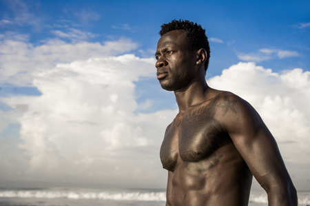 close up head and shoulders portrait of young attractive and fit black African American man with strong muscular body posing cool model attitude on the beach isolated on Summer blue skyの写真素材