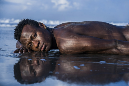 artistic expressive portrait of young attractive and sexy black afro American man with athletic muscular body posing cool in sea water on desert beach in male beauty and plasticity conceptの写真素材