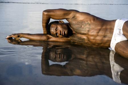 artistic expressive portrait of young attractive and sexy black afro American man with athletic muscular body posing cool in sea water on desert beach in male beauty and plasticity conceptの写真素材