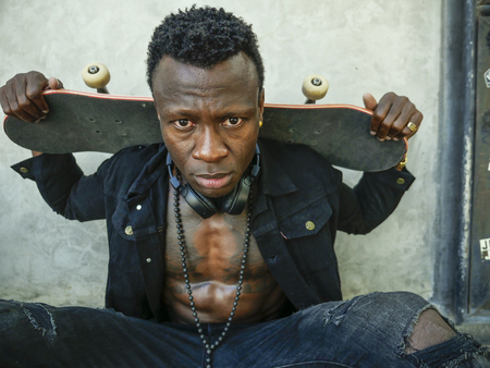 cool isolated portrait of young attractive and confident hipster black afro American man leaning on street wall holding skate board as badass guy in urban style conceptの写真素材
