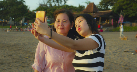 young happy and pretty Asian Chinese girl on the beach taking selfie photo with her mother, a 60s mature woman , enjoying Summer holidays travel together at beautiful tropical destinationの写真素材