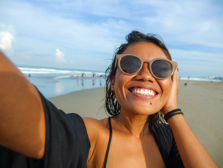 young happy and attractive Asian exotic woman on the beach taking selfie portrait with mobile phone smiling cheerful enjoying holidays trip at beautiful tropical island in social media lifestyleの写真素材