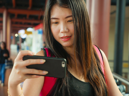lifestyle portrait of young happy and beautiful Asian Korean or Japanese woman taking selfie picture with mobile phone at airport terminal in travel social media blogger and holidays trip conceptの写真素材