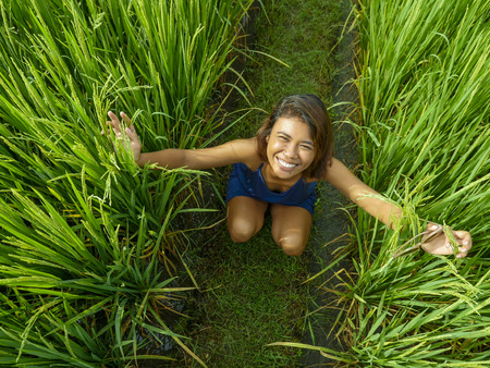 natural and fresh portrait of young happy and exotic islander Asian girl from Indonesia smiling cheerful and excited posing in green field playing with rice enjoying holidays tripの写真素材