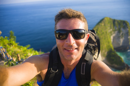 young happy and attractive backpacker man taking selfie portrait photo with mobile phone in front of amazing sea landscape smiling cheerful feeling free trekking on beautiful beach cliffの写真素材