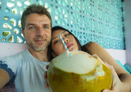 natural lifestyle portrait of young beautiful and happy mixed ethnicity couple in love relaxed together with handsome Caucasian man and beautiful Asian Korean woman drinking healthy coconut waterの写真素材