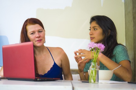 lifestyle portrait of two young happy and beautiful caucasian and latin women working at office cafe with laptop computer discussing as digital business partners and diversity ethnicity friendsの写真素材