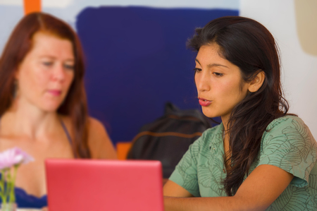 lifestyle portrait of two young happy and beautiful caucasian and latin women working at office cafe with laptop computer discussing as digital business partners and diversity ethnicity friendsの写真素材