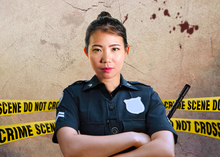 young Asian American police officer standing serious in custody of crime scene for preserving evidence at do not cross police line yellow tape in murder investigation and law enforcement conceptの写真素材