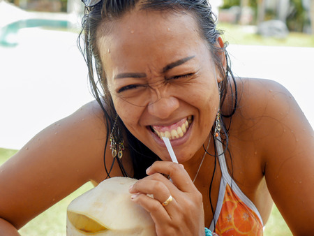 lifestyle natural portrait of 30s or 40s happy and attractive Asian Indonesian woman in bikini drinking coconut water at tropical resort swimming pool smiling cheerful and relaxed enjoying holidaysの写真素材