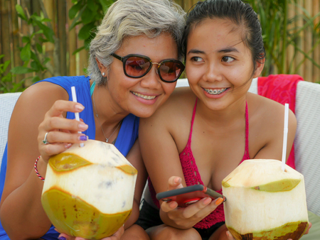 happy and beautiful Asian Indonesian teenager girl using mobile phone enjoying Summer holidays drinking coconut at tropical resort garden together with her mother, an attractive 40s womanの写真素材