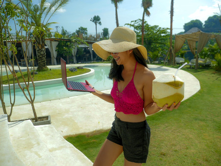 young beautiful and happy Asian Indonesian teenager woman in bikini at tropical resort swimming pool networking relaxed and chilled with laptop computer smiling enjoying Summer holidaysの写真素材