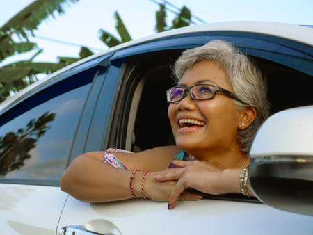 lifestyle portrait of attractive and happy middle aged Asian Indonesian woman 40s or 50s  with grey hair and beautiful smile sitting in her car enjoying nature looking through window relaxed and cheerfulの写真素材